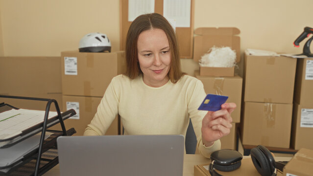 Woman holding creditcard and typing on laptop in packing warehouse among stacked parcels and barcode scanner; entrepreneurial focus.