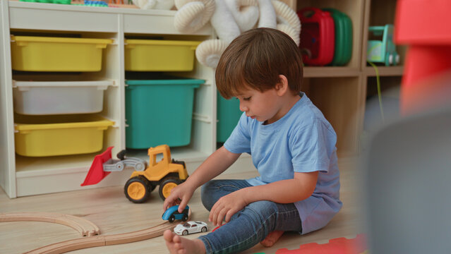 Boy sitting on floor playing with yellow toy tractor and wooden tracks in a building playroom, reaching for a small car; focused curiosity.