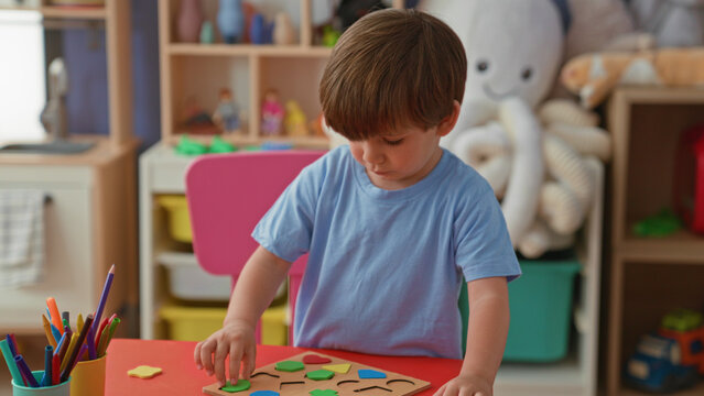 Boy placing wooden shape puzzle pieces with hands at red table in building; concentration learning play.