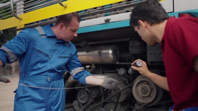 Two mechanics work side by side on a large vehicle engine. One man in blue overalls points at engine parts while the other holds a flashlight