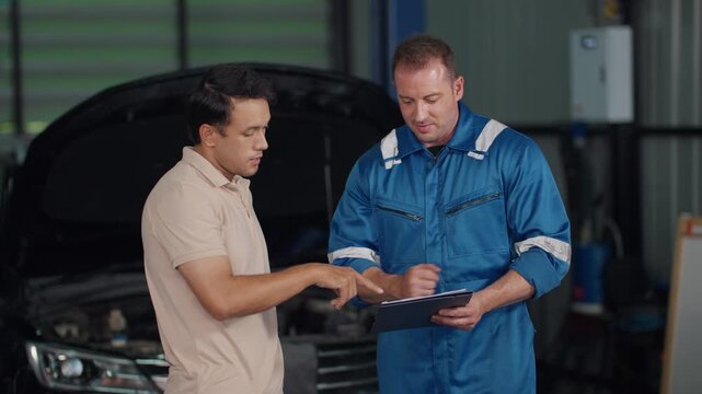 Mechanic explains vehicle issue to customer. Customer listens intently, hands on hips. Mechanic holds tablet, gestures toward engine