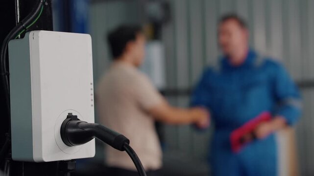 White charger plugs into industrial wall outlet. Two men shake hands near charging station. One holds red folder, other wears blue uniform