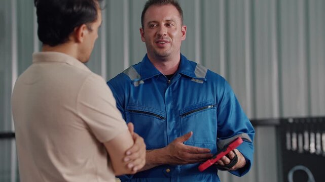 Two men converse in a workshop. One wears a blue work uniform, holding a tablet. The other, in a beige shirt, listens attentively