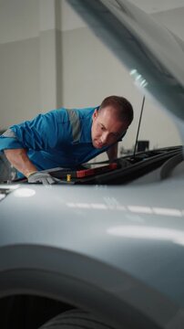 Focused mechanic examines car engine under open hood. He wears blue uniform, gloves, leans over silver vehicle. Workshop background shows tools