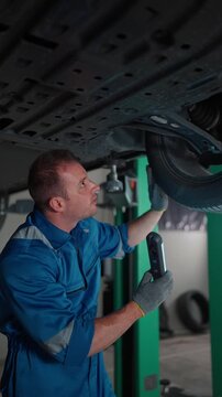 Mechanic inspects car underside with flashlight and tool. Focused expression shows careful attention to detail. Ideal for automotive repair