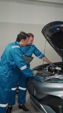 Two mechanics in blue uniforms stand beside a silver car. They examine engine components under an open hood. One holds a tablet