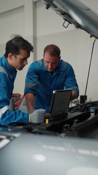Two mechanics in blue uniforms work together. They examine car engine components under open hood. One uses laptop while other observes closely