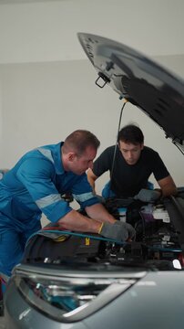 Two mechanics work under a car's open hood. One smiles, gesturing while explaining. The other listens, focused on the engine. They are in a clean