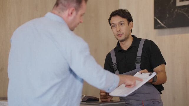 Two men discuss work in a cozy office setting. One man, wearing overalls, writes on a clipboard. The other, in a light shirt, listens attentively