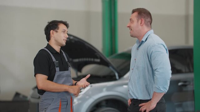 Two men discuss car issues in a garage. One wears overalls, the other in a shirt. They stand near an open hooded vehicle