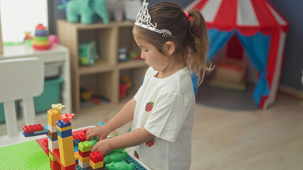 Girl with tiara holds green toy frog and stacks colorful plastic building blocks, playing at a play table inside a building  playful curiosity. © Krakenimages.com