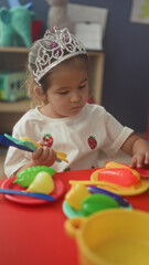 Girl wearing tiara sits at red table and uses hand to pick plastic toy food and utensils in building  quiet concentration. © Krakenimages.com