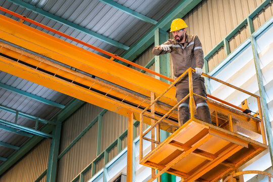 Industrial technician in hard hat inspecting overhead crane structure from platform, Maintenance engineer standing on aerial basket platform pointing during factory inspection