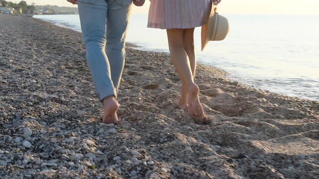 Barefoot couple walking on pebble beach, legs of man in jeans and woman with straw hat strolling by sea at sunset. Summer vacation and romantic walk concept, low angle