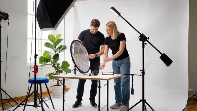 Photographer trains an assistant to position a reflector and studio lights around a tabletop setup during a practical photography studio session