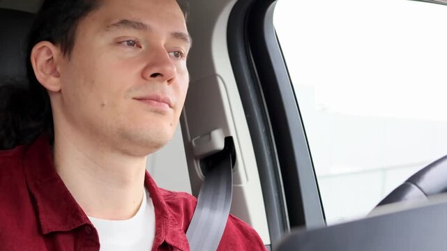 Close-up of a smiling man in a burgundy shirt with a fastened seatbelt nodding his head to the rhythm of music while driving through the city. Concept of a good mood and enjoying the morning commute.