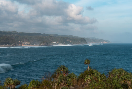 Scenic wide view of deep blue ocean waves hitting rocky tropical coastline with green hills and cloudy sky
