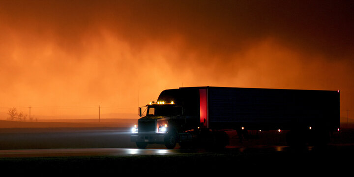 A large semi-trailer truck traveling on a highway at night, illuminated by its headlights and the glow of a distant fire or sunset