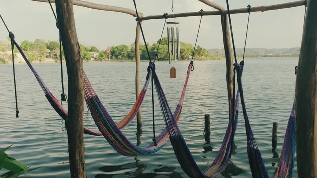 Colorful hammock beds sway gently over tranquil water at a peaceful lakeside retreat. Natural wood frame, calm Lake Peten Itza, lush greenery, and warm daylight add to the serene vibe.
