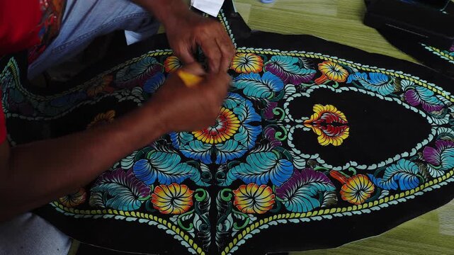 Close-up of hands meticulously painting a vibrant, floral pattern onto a black fabric, likely a kite or traditional textile