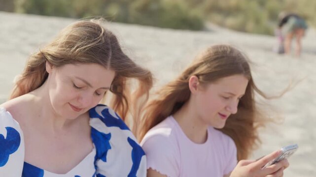 A girl is sitting on the beach with phone slow motion next to her sad mother camera movement medium shot
