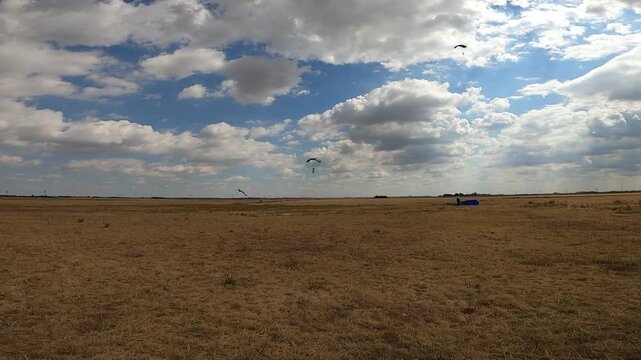 Skydivers using wing-type parachutes land under a beautiful, cloudy sky in strong winds