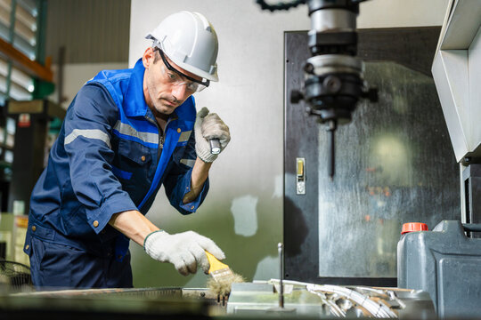 Industrial engineer using flashlight and brush to inspect and clean CNC machine part, Factory technician cleaning CNC equipment with paintbrush while holding flashlight
