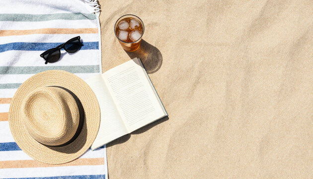 Summer beach flat lay with striped towel, sunglasses, straw hat, book and iced drink on sand, top view