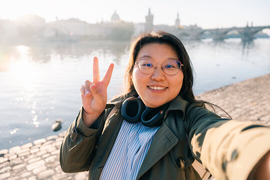 Korean tourist takes selfie by river in Prague, peace sign gesture