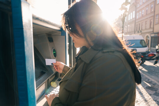 Woman using ATM while holding card in sunny urban setting at daytime