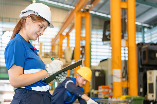 Female industrial engineer holding clipboard and taking notes in manufacturing factory, Factory supervisor writing notes with technician working in background