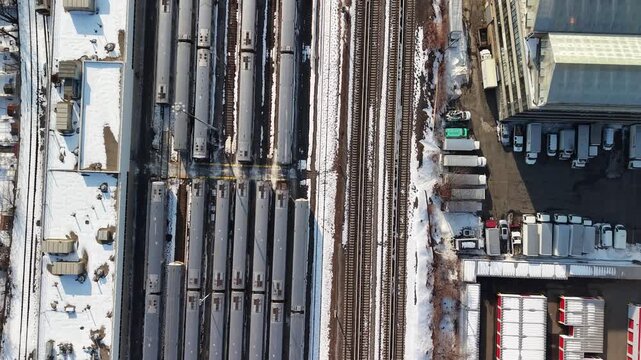 Top down drone footage directly over a snow covered rail yard in Queens, New York City, parallel tracks and rows of parked train cars filling the frame, a parking lot and industrial yards at the edges