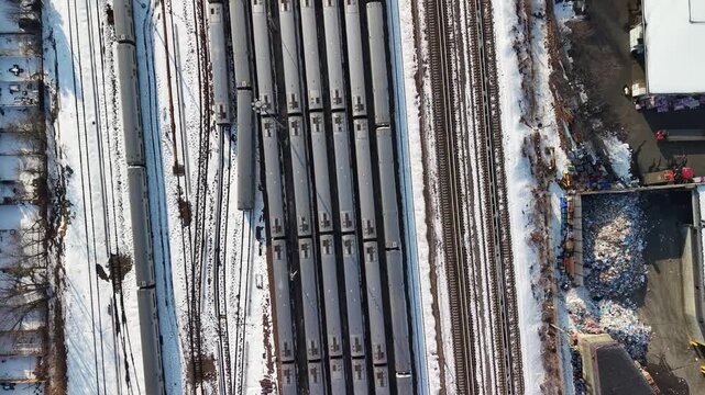 Top down drone footage directly over a snow covered rail yard in Queens, New York City, parallel tracks and rows of parked train cars filling the frame, a parking lot and industrial yards at the edges