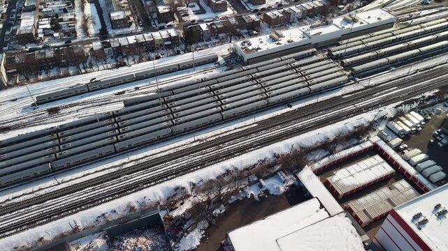 Aerial drone footage over a snow covered rail yard in Queens, New York City, long rows of parked silver commuter trains on parallel tracks, industrial buildings and homes around the yard