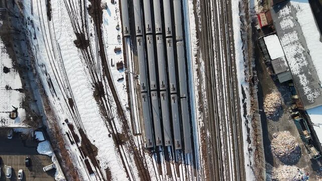 Top down drone footage directly over a snow covered rail yard in Queens, New York City, parallel tracks and rows of parked train cars filling the frame, a parking lot and industrial yards at the edges