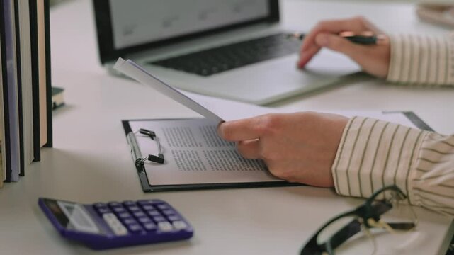 Person reviewing printed financial documents and writing notes beside calculator, laptop, and eyeglasses at a home office desk. checking financial figures for accounting analysis and business report p