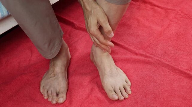 Close-up of an elderly man massaging his ankle with his hand; he is sitting on his bed at home with his feet resting on the floor