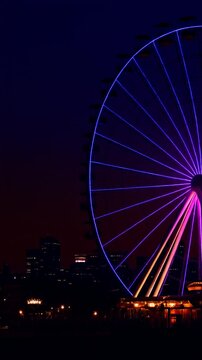 Large Ferris wheel with color changing LED lights glowing against a dark city skyline at night, red orange purple and blue illumination, vertical video