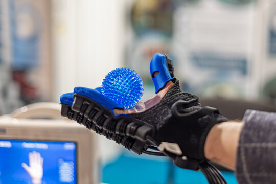 Close up of a robotic rehabilitation glove holding a blue spiky massage ball during hand therapy, innovative medical device designed to assist finger movement and motor recovery.