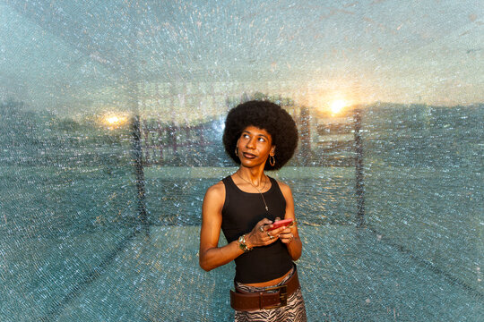 Non binary person holds smartphone behind broken glass at sunset