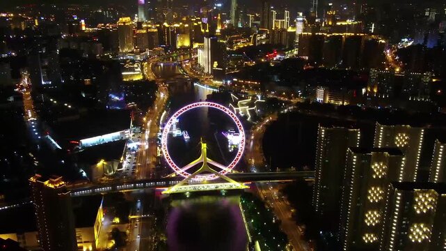 Aerial drone view of the brightly lit Tianjin Eye Ferris wheel straddling the Hai River at night, surrounded by illuminated skyscrapers, active road traffic, and the vibrant downtown Tianjin skyline