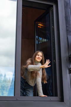 Young woman in gray sweater leaning out of an open window and waving her hand with a smile, cheerful girl enjoying fresh air at a modern mountain cabin.