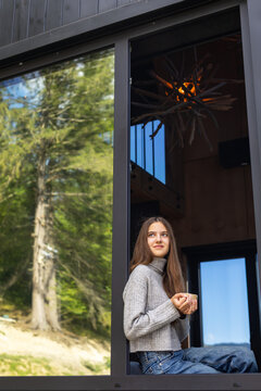 Young woman in gray sweater sitting by a large open window with a cup and looking outside, peaceful relaxation in a modern forest cabin with scenic woodland views.