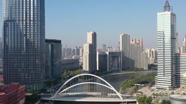 Aerial drone view of Tianjin&rsquo;s Hai River waterfront with modern glass skyscrapers, the distinctive white Binjiang Bridge, and light traffic under a clear blue daytime sky