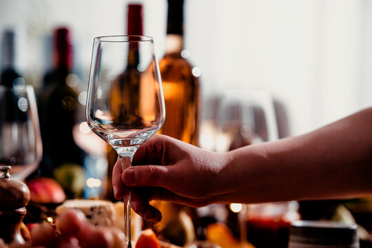Hand holding empty wine glass at festive lunch table, autumn celebration setup. Luxury hospitality service concept. Elegant crystal glassware with blurred wine bottles in warm background.