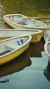 cinematic vertical shot panning right across vintage pastel yellow and blue rowboats gently floating on green rippling water in a quiet nostalgic park pond