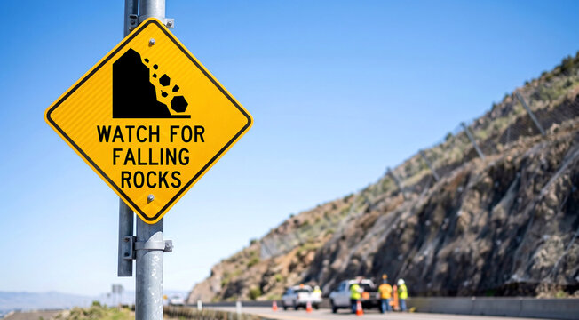 Yellow road sign on highway beside rocky slope with road crew and cones, rockfall hazard and highway safety concept