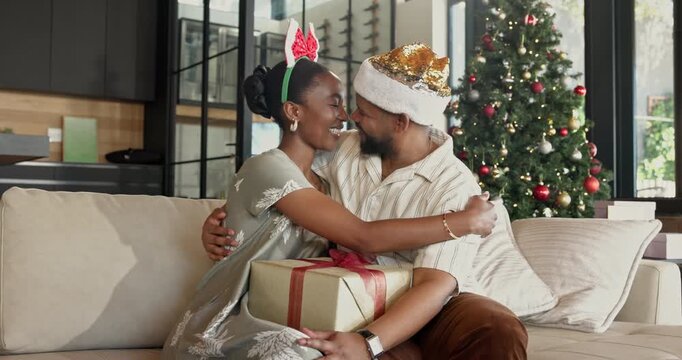 Christmas, gift and black couple kiss on home sofa for bonding, quality time and gratitude. Happy man, woman or festive season in house with hug, xmas present and surprise for holiday celebration