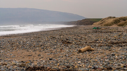A wide pebble beach with scattered driftwood, rolling waves, and distant coastal cliffs under soft, hazy light near Llanengan on the Llŷn Peninsula. Natural coastal scene with strong textures and an u © Bernd Brueggemann
