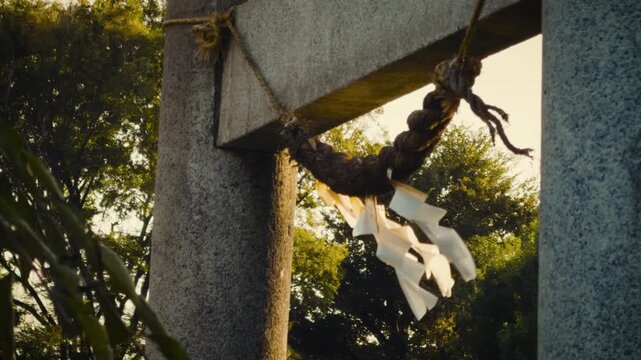 cinematic horizontal low angle shot panning right on a traditional japanese stone torii gate with a sacred shimenawa rope and white shide paper streamers blowing in the wind during golden hour sunset